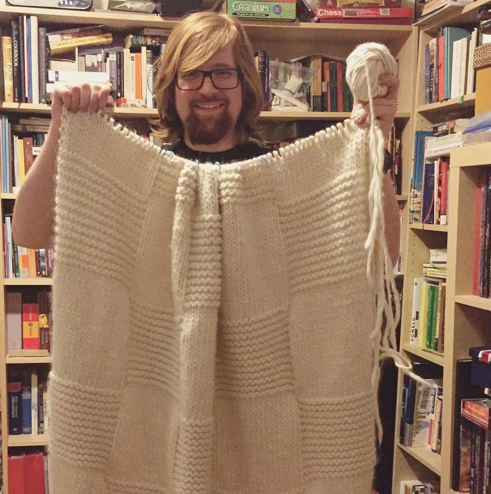 A photo of Leif holding up a giant knitted blanket that he made in 2018.